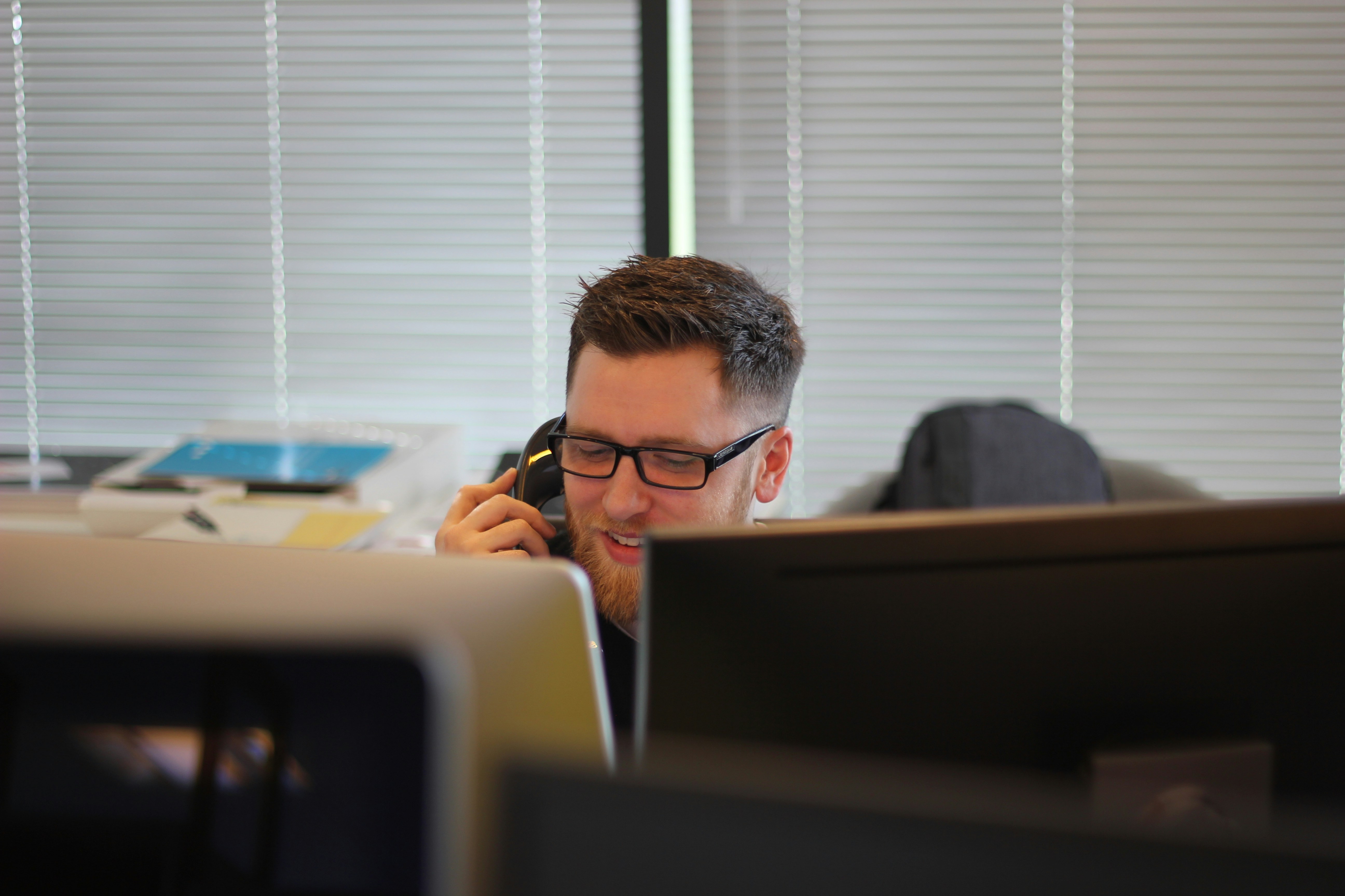 man with glasses talking on the phone at his desk in a office discussing debt recovery agencies with a debt collector