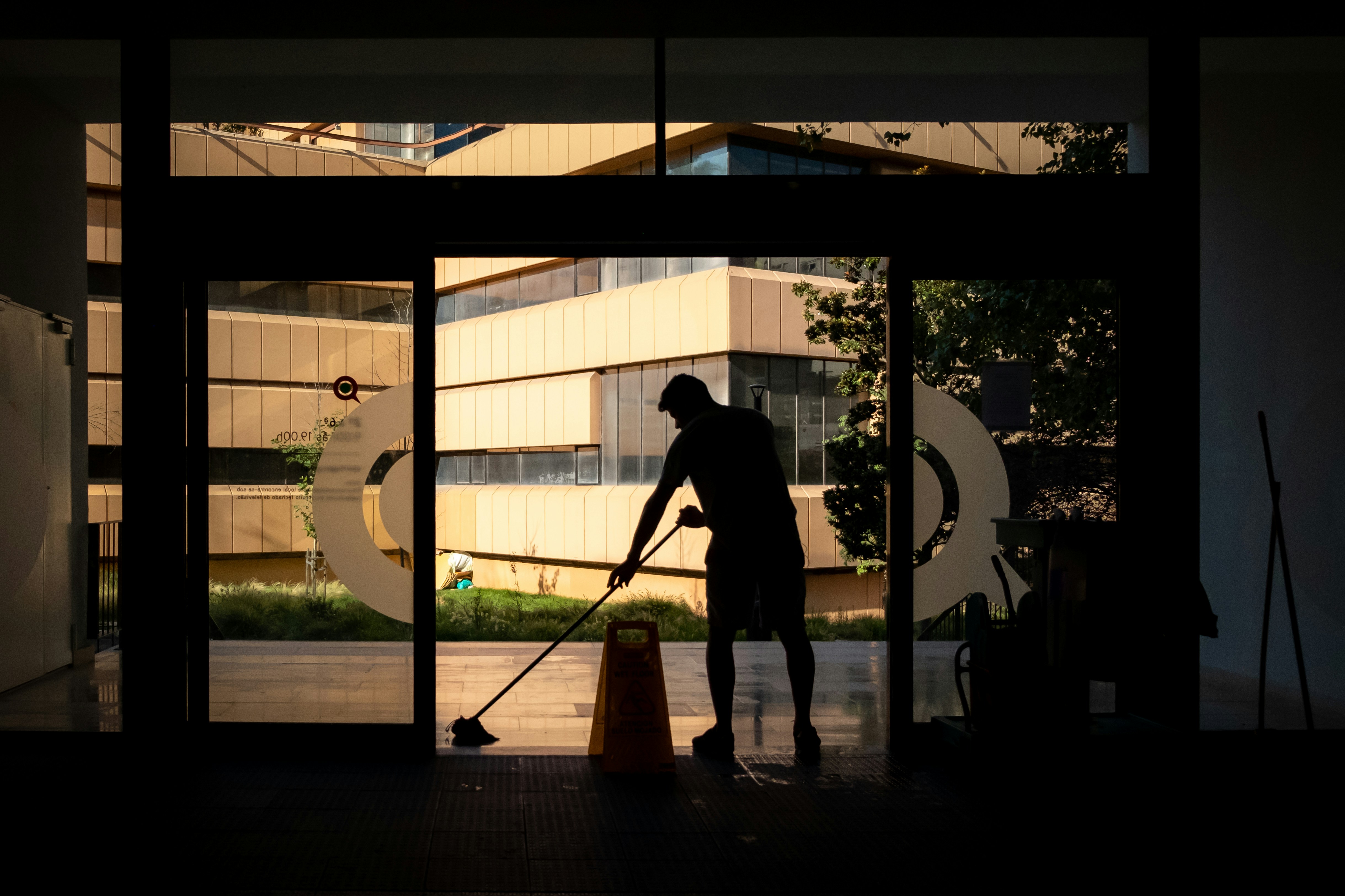 man moping the floor of a commercial building with the glass doors open and a wet floor sign